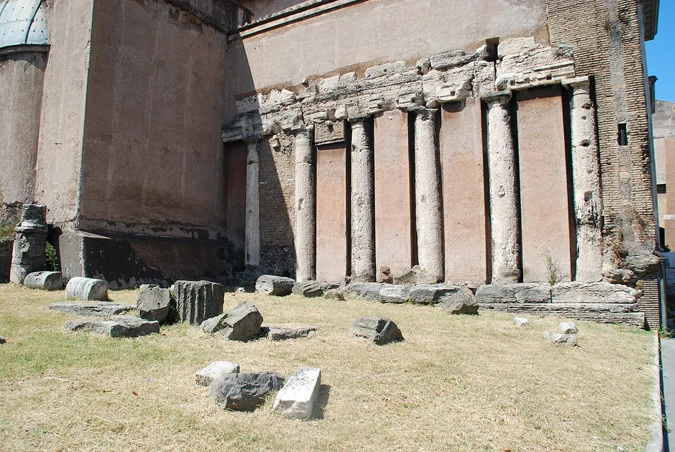A row of weathered stone columns and a broken entablature built into the plastered side wall of a church, with column drums and blocks lying on dry grass in front.