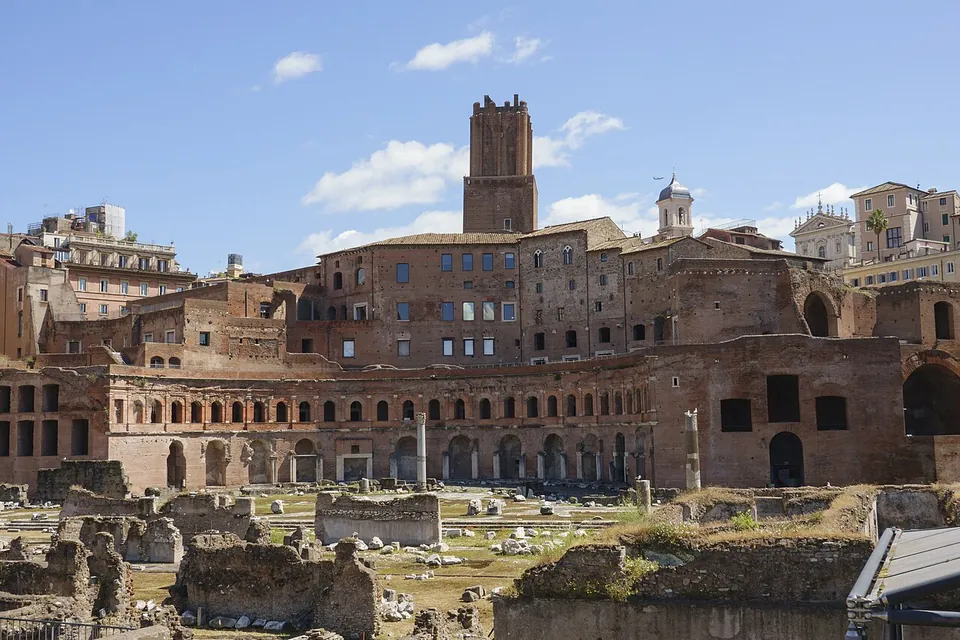 Low excavated walls, column stumps and scattered blocks on grass in the foreground, and behind them a tall curved brick building of several storeys with rows of arched openings, a square brick tower rising above it under a blue sky.