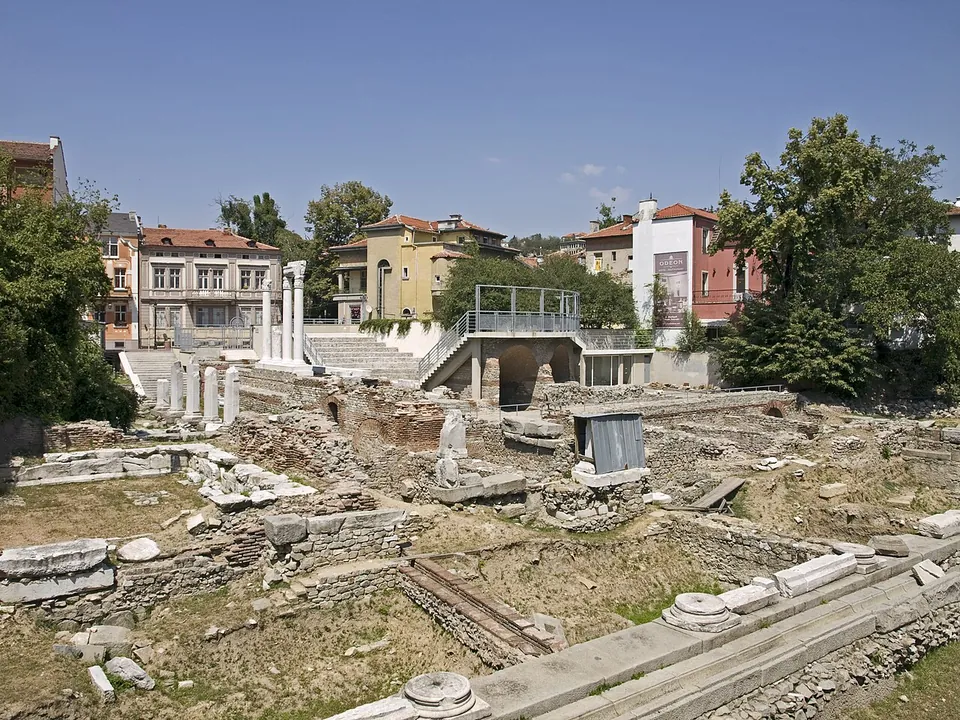 Excavated stone foundations, column bases and fallen blocks of the ancient forum spread across a sunken area of grass, with a few standing columns on the left and the painted houses of modern Plovdiv above it.