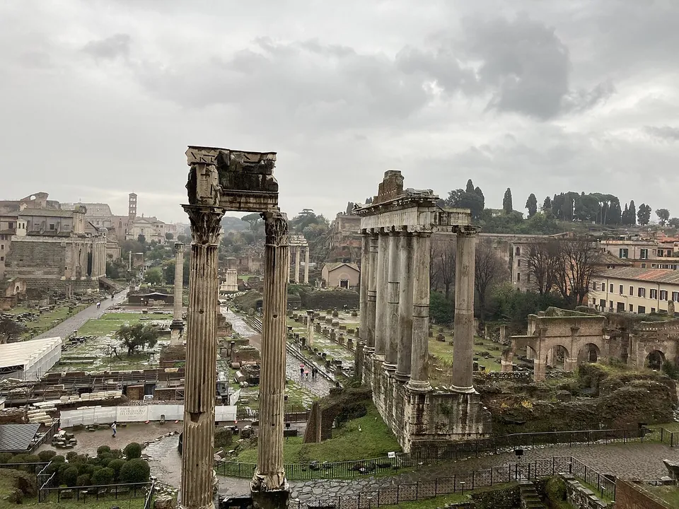The length of the Forum seen from above at its western end under a grey sky, three tall columns carrying a fragment of entablature in the foreground and a row of taller columns on a podium to the right, with paths, ruins and trees stretching away and the Colosseum on the horizon at the left.