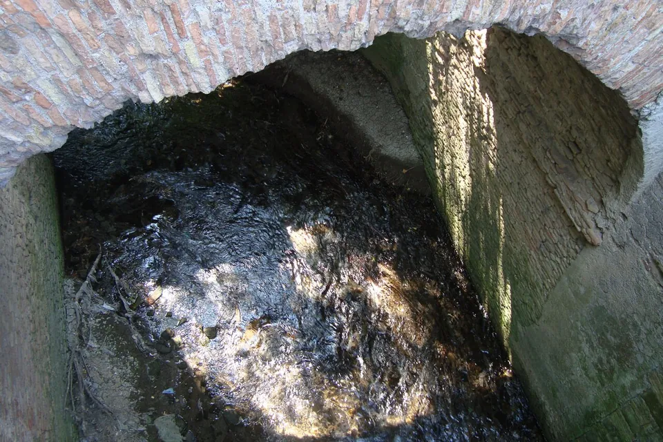 Looking down at a dark stream flowing under a low brick arch, between walls of brick and stone patched with moss.