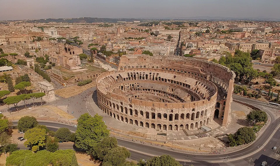 An aerial view of the whole oval amphitheatre. Along one side the outer wall stands up to its top storey; along the other it has broken away, leaving a lower ring of arches. Inside, a grid of brick walls fills the arena, and roads, trees and the roofs of the city surround the building.