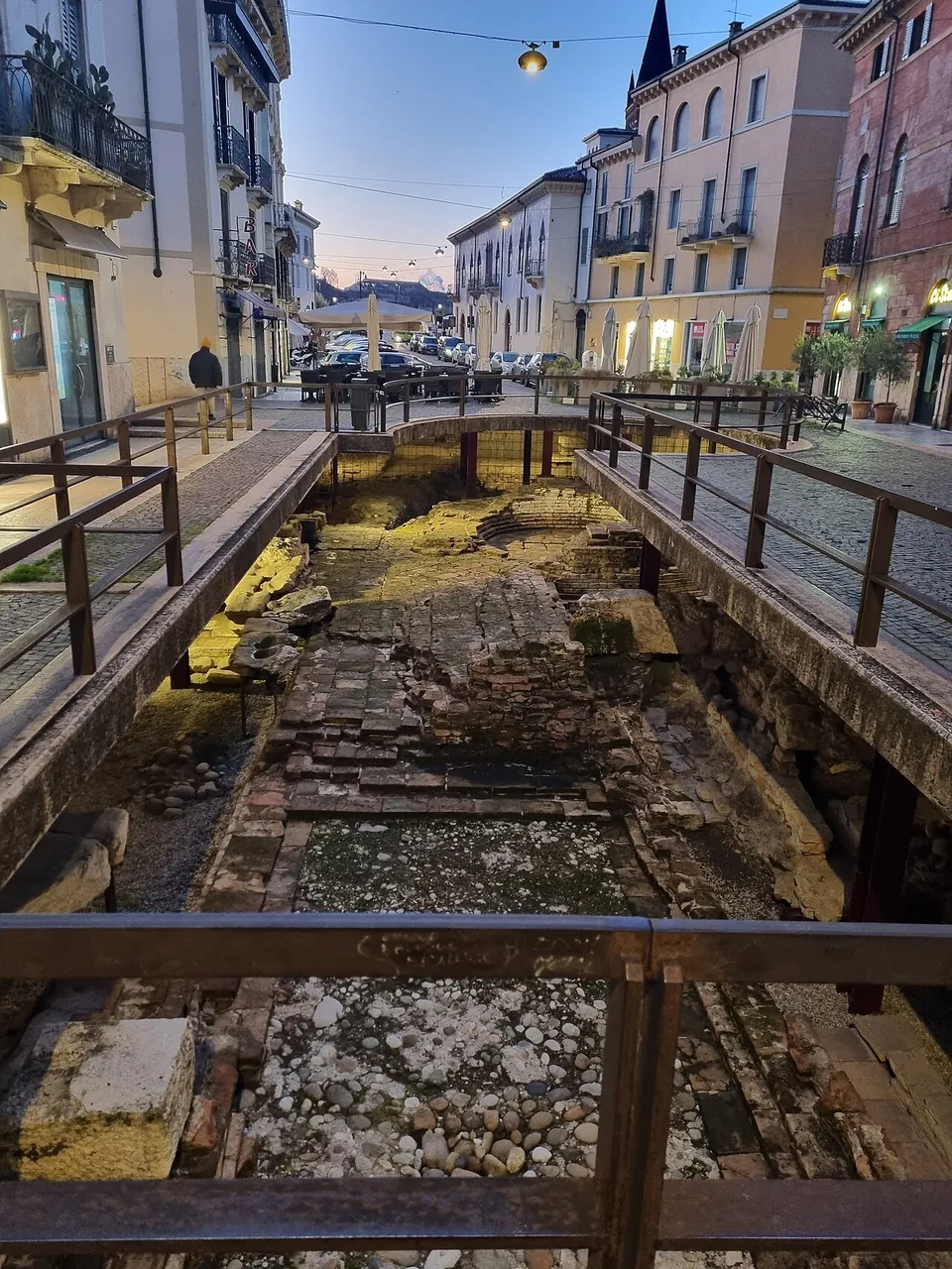 A long excavation trench lit at dusk in the middle of a street, between iron railings, its floor of brick and stone foundations, with houses and parked scooters beyond.