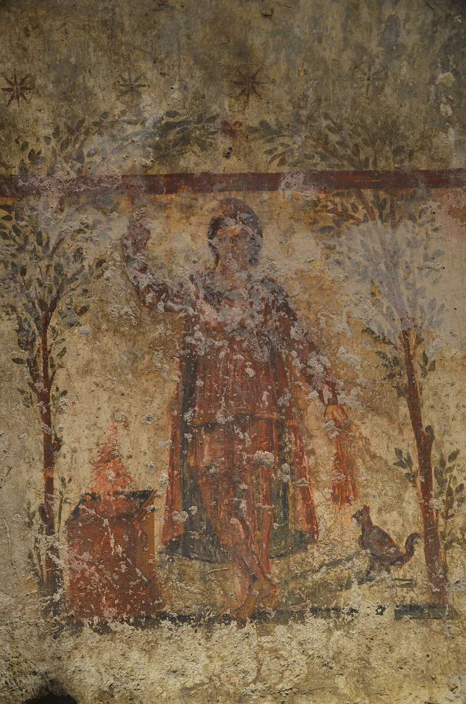 A faded wall painting of a standing figure in a long red garment holding up a torch, between two slender trees, with a small altar at the left and a small animal at the right, under a band of red and a ceiling scattered with painted stars.