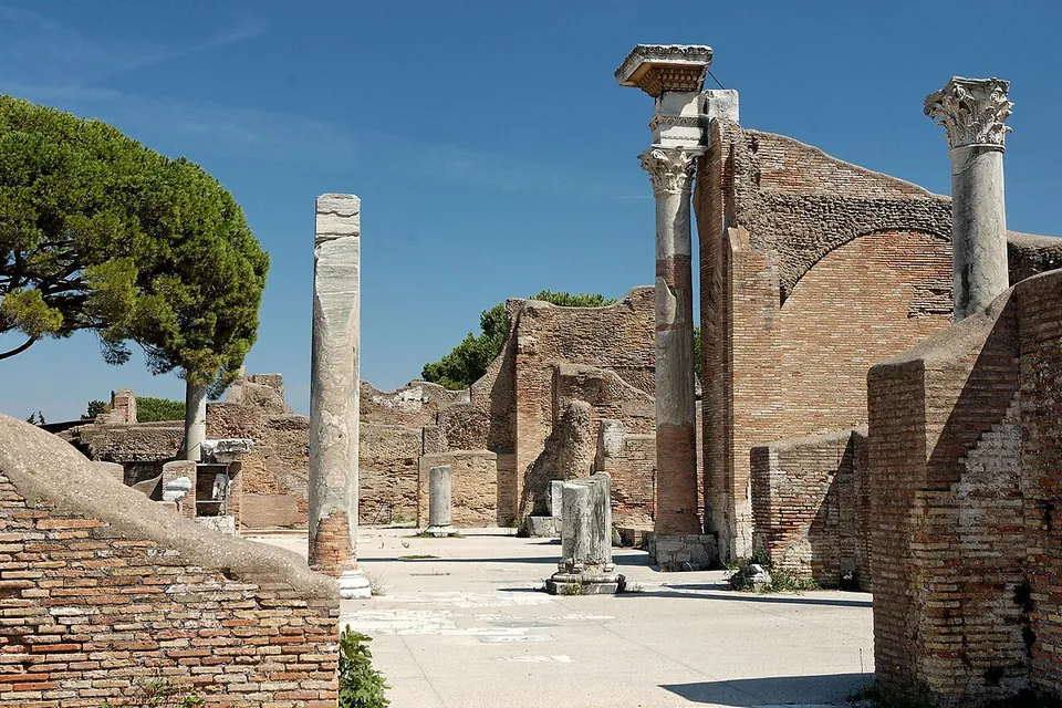 A paved hall between high walls of red brick, with a broad brick arch behind and tall marble columns standing on the floor, two of them still carrying their carved capitals, against a blue sky.