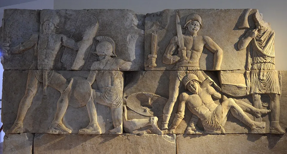 A long stone relief in high relief showing gladiators in combat, with helmets, shields and swords, one fallen figure lying across the lower register and a robed figure standing at the right.