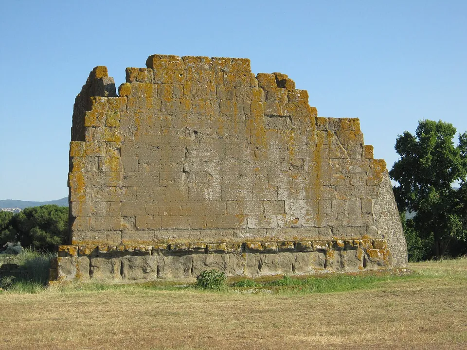 A tall free-standing wall of squared, lichen-spotted stone blocks, ragged along its top edge, rising from a low stepped base in a mown field, with trees and distant hills behind.