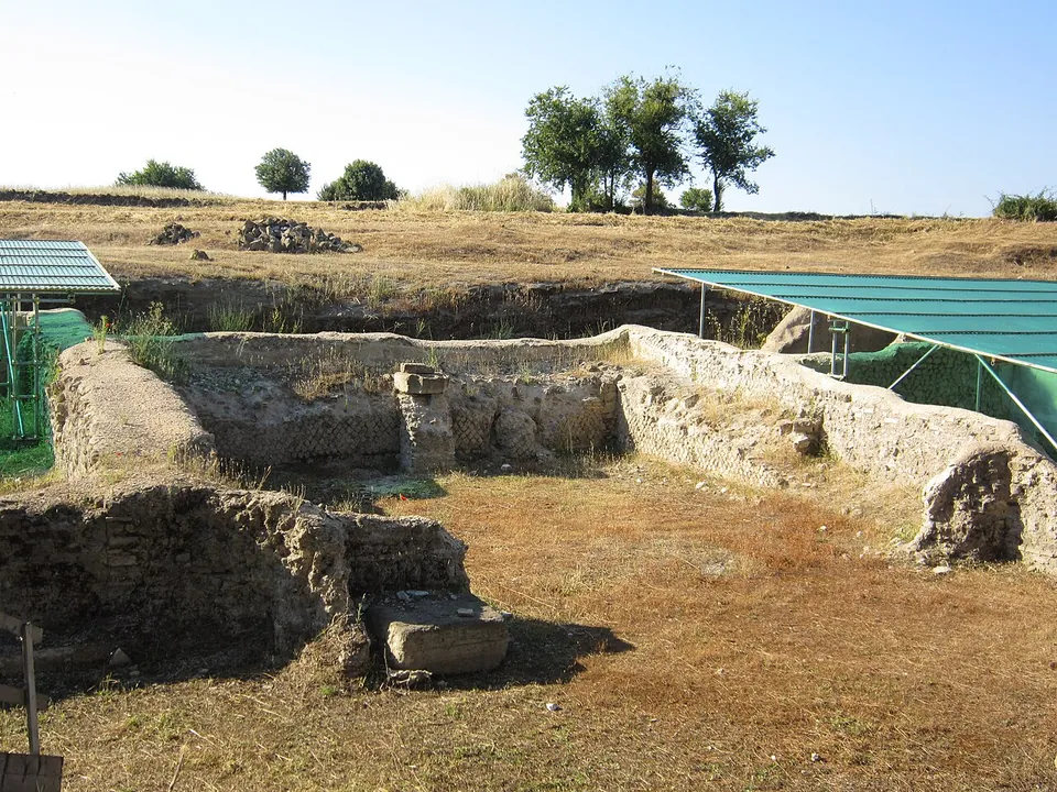 Low walls of rubble and squared stone laid bare in a shallow excavation, with green shade roofs on frames over the neighbouring trenches and trees on the field edge beyond.