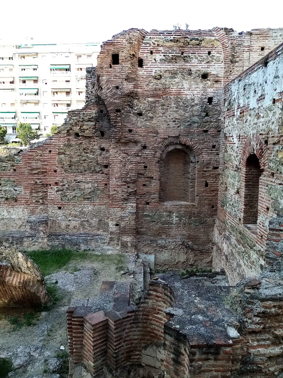 A high ruined wall of red brick bands and stone, with an arched niche and rows of small square holes, above the brick footings of a room.