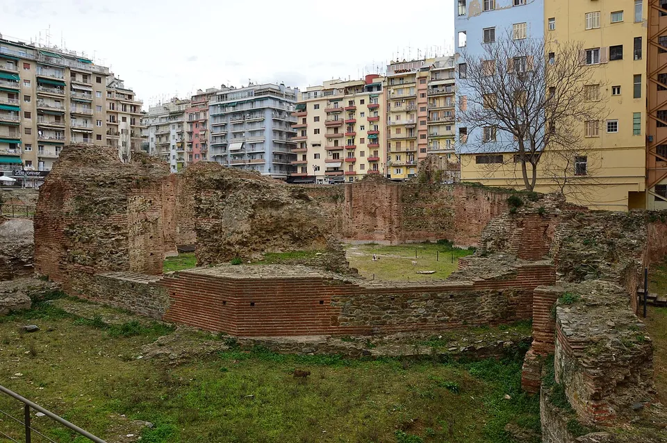Ruined walls of thin red bricks and rubble enclosing an eight-sided hall with a grassy floor, sunk below street level among tall apartment blocks.