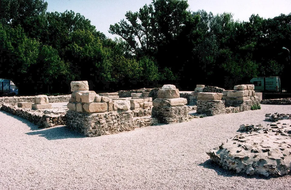 Stone piers built of large squared blocks standing on footings of rough rubble masonry around a gravel courtyard, with a broken stretch of rubble wall in the foreground and dense trees behind.