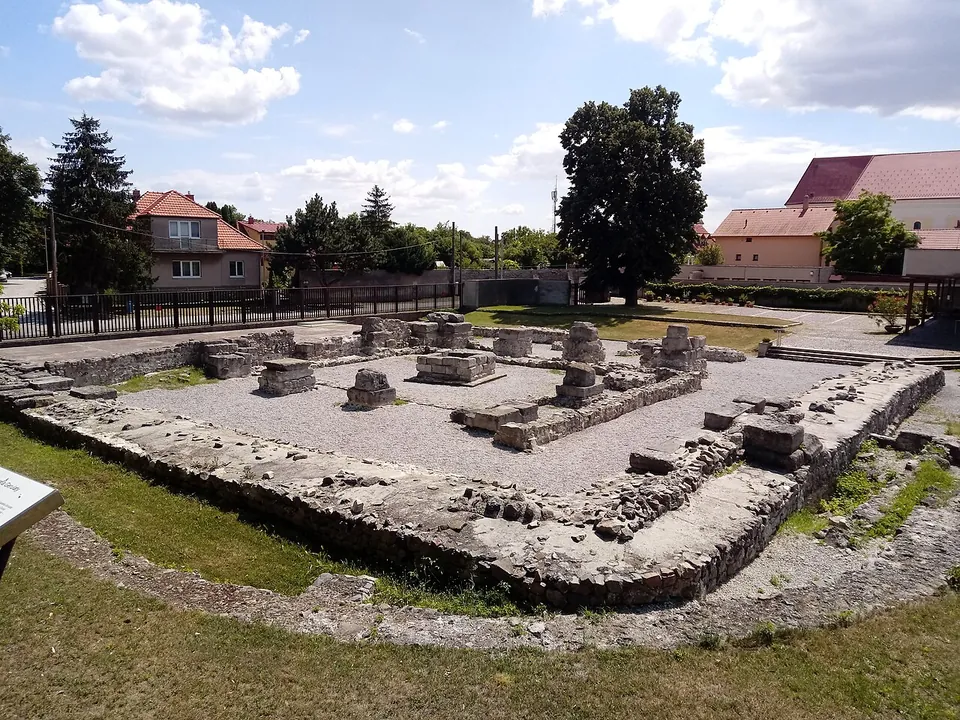 A square enclosure of low, grassy-topped stone walls around a gravel courtyard, with a row of stone piers and blocks set out inside it; houses with red roofs, a fence and tall trees surround the site.