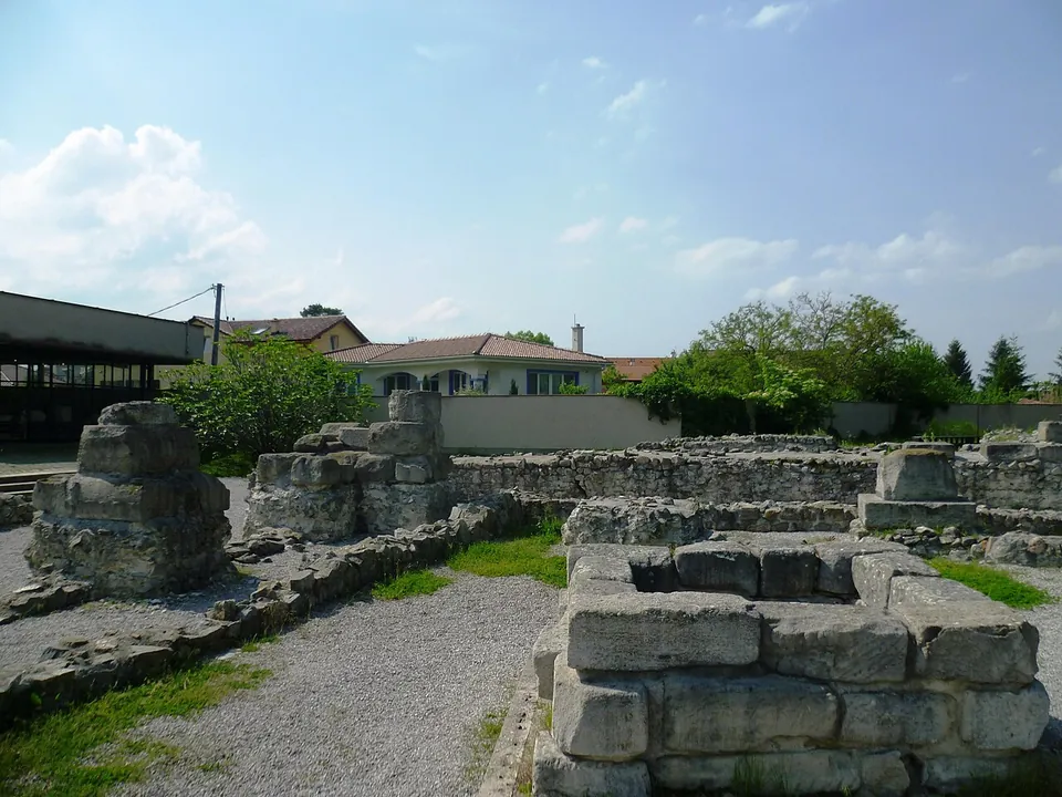 A pier of large, weathered squared stone blocks in the foreground, with low rubble walls and gravel beyond, a flat-roofed modern building on the left and family houses behind.