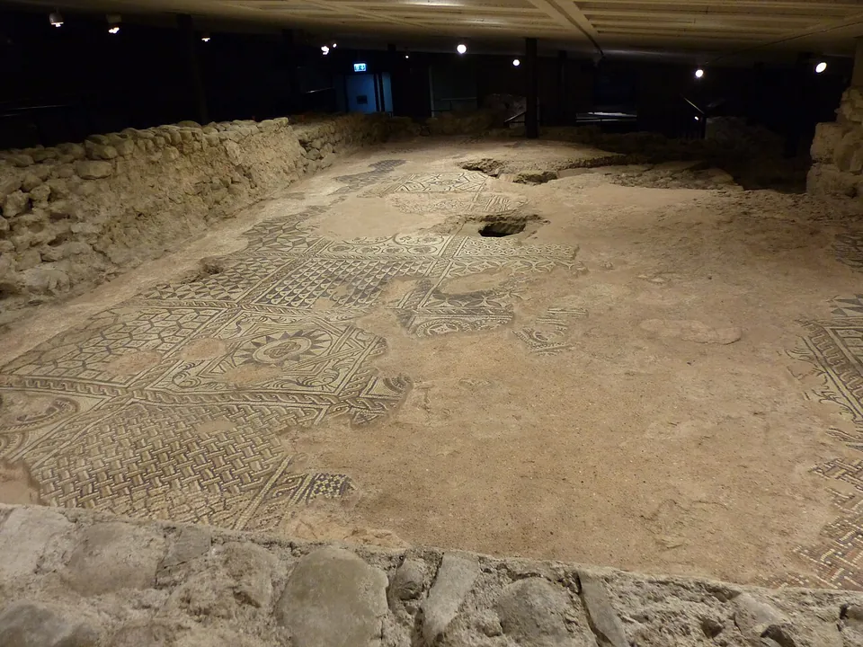 A long mosaic floor in a dim underground hall, laid in geometric panels of interlace, chequers and a rosette in dark and pale stones, patchy and worn in places, with a low rubble wall along one side.
