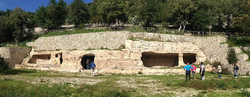 A low limestone face below a stone retaining wall, cut with several rectangular openings into rock-cut rooms, with visitors on the grass in front.