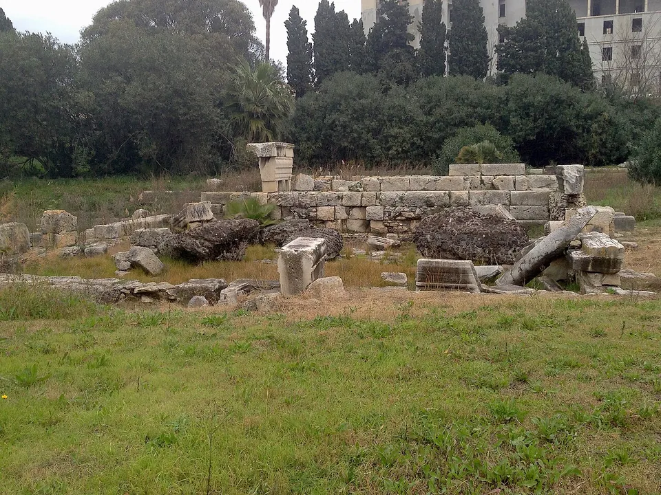 Low walls of squared stone blocks, a fallen column shaft and loose blocks on a grassy plot, with cypresses and a modern apartment building behind.