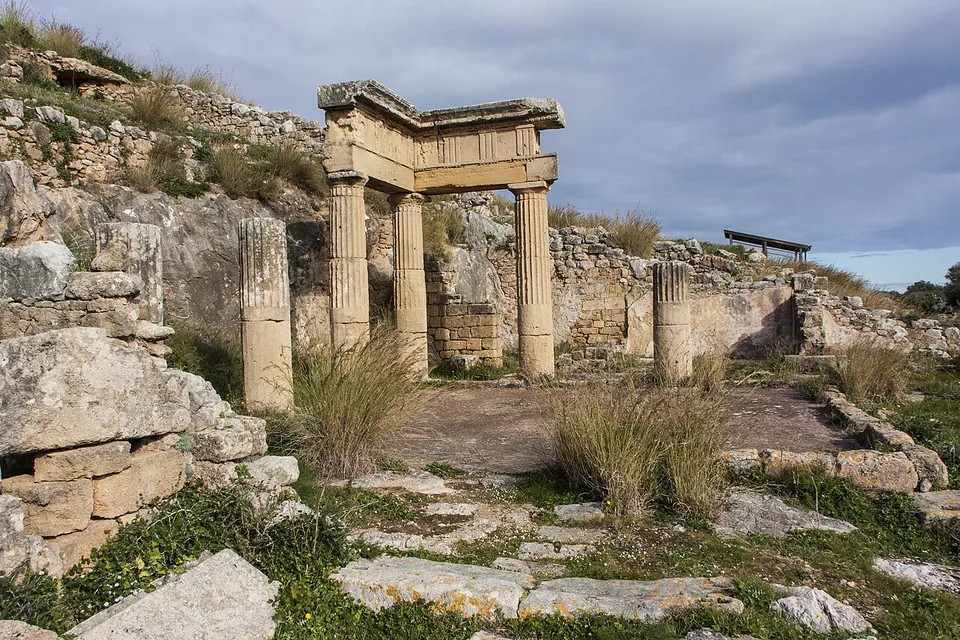 Three fluted stone columns carrying a corner of entablature, with further column shafts standing on their own, round a paved courtyard among ruined walls on a hillside.