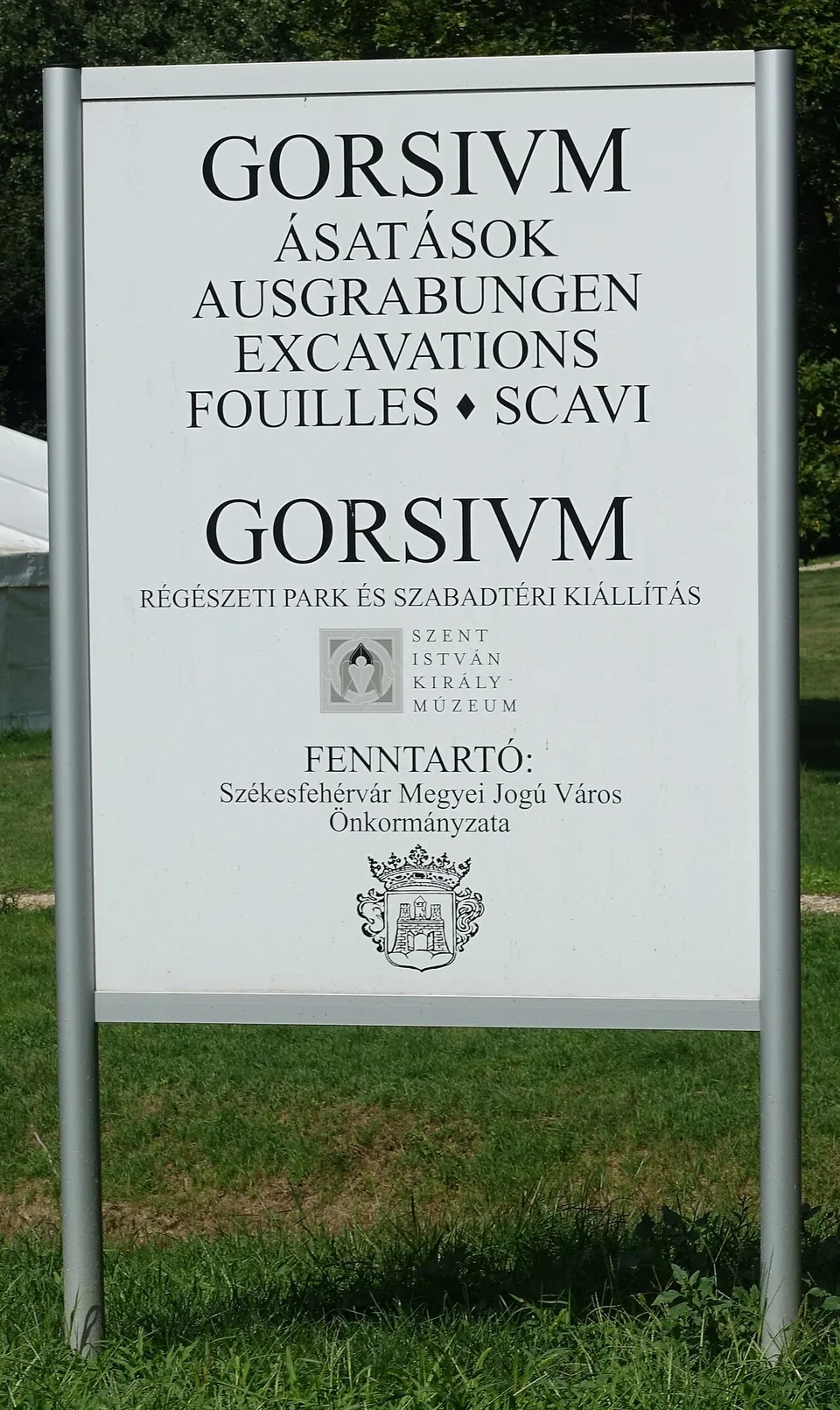 A white sign on two posts reading "GORSIVM - ásatások, Ausgrabungen, Excavations, Fouilles, Scavi - Régészeti park és szabadtéri kiállítás", with the logo of the Szent István Király Múzeum and, as maintainer, the city of Székesfehérvár.