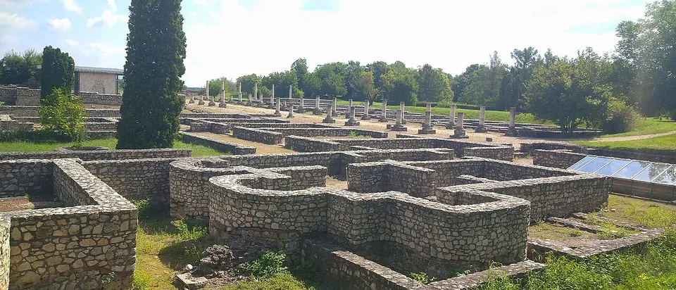 A maze of conserved stone foundation walls in the foreground, with a long row of stone column bases standing on a lawn behind them and a tall cypress on the left.