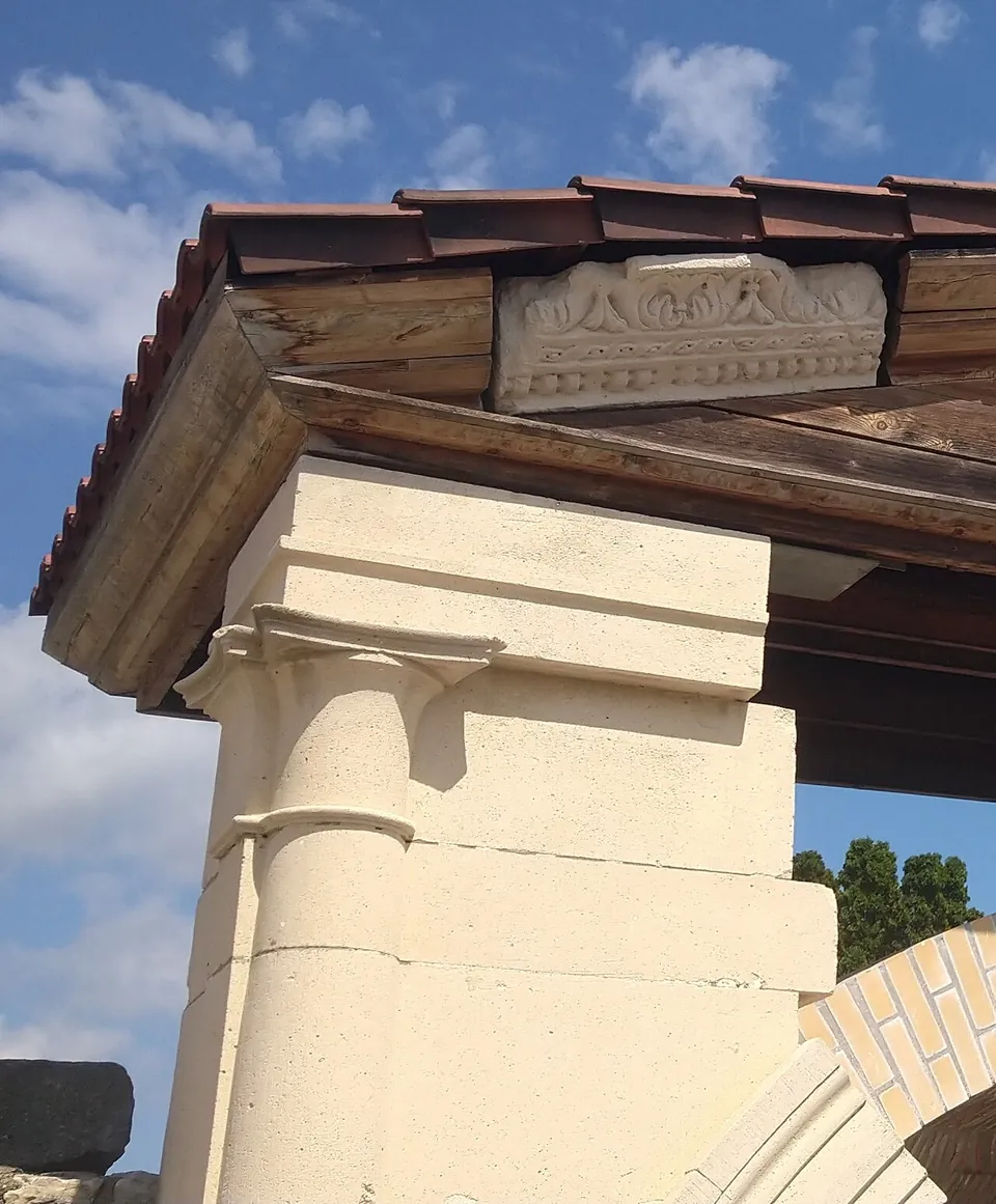 The corner of a pale stone structure with a moulded pilaster capital, under a timber and tiled roof whose gable holds a carved stone panel decorated with leaves.