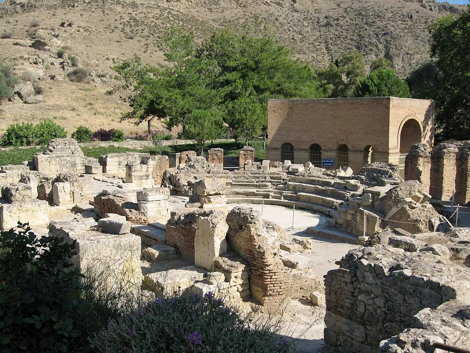 The curved stone seating of a small Roman theatre among ruined walls of stone and brick, with a modern brick building with arched windows beside it and a bare hillside behind.