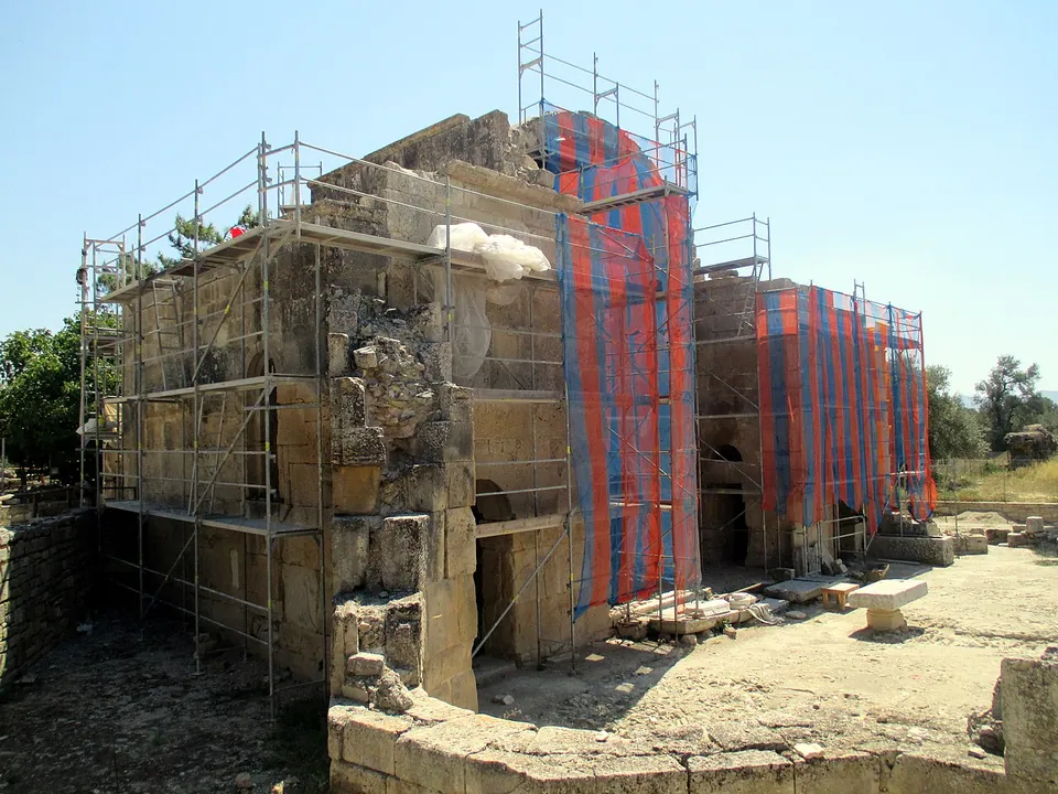 The tall stone walls of a ruined church apse, wrapped in scaffolding and coloured protective netting, beside open excavated ground.