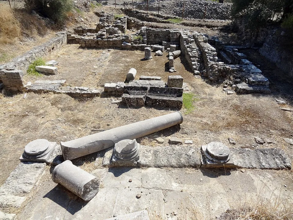 Low walls of rough stone and a few squared blocks rising among dry ground and scrub, with olive trees and a hillside behind.