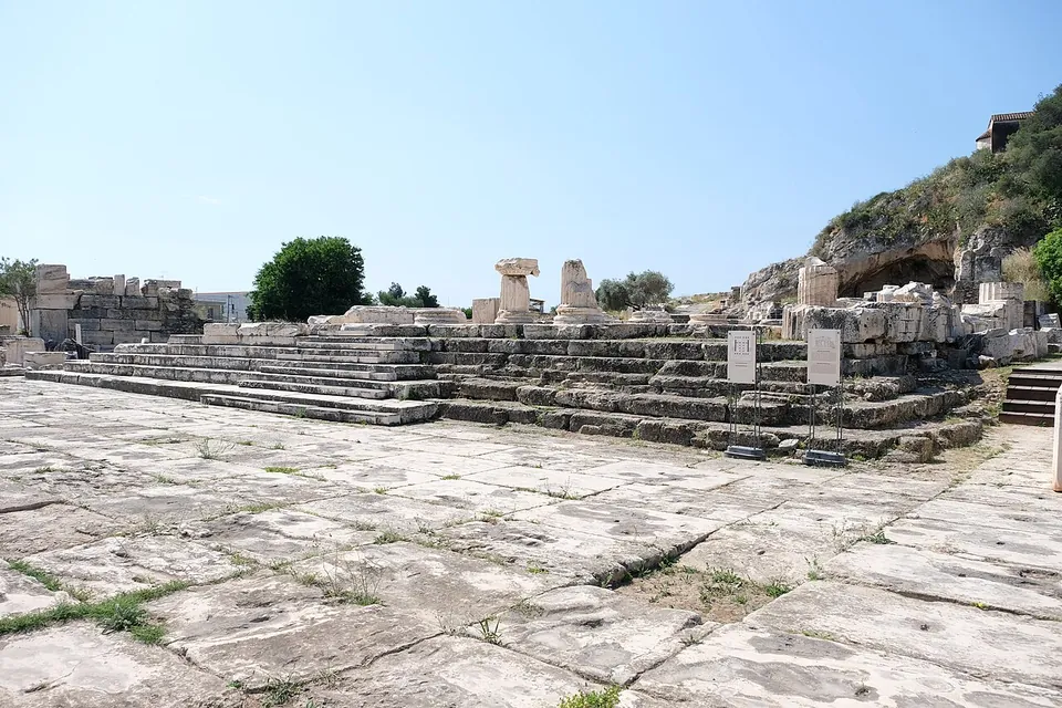 A broad flight of marble steps rising from a wide paved forecourt to the stumps of columns and piled blocks of a ruined gateway, with a rocky hillside and a cave mouth on the right.
