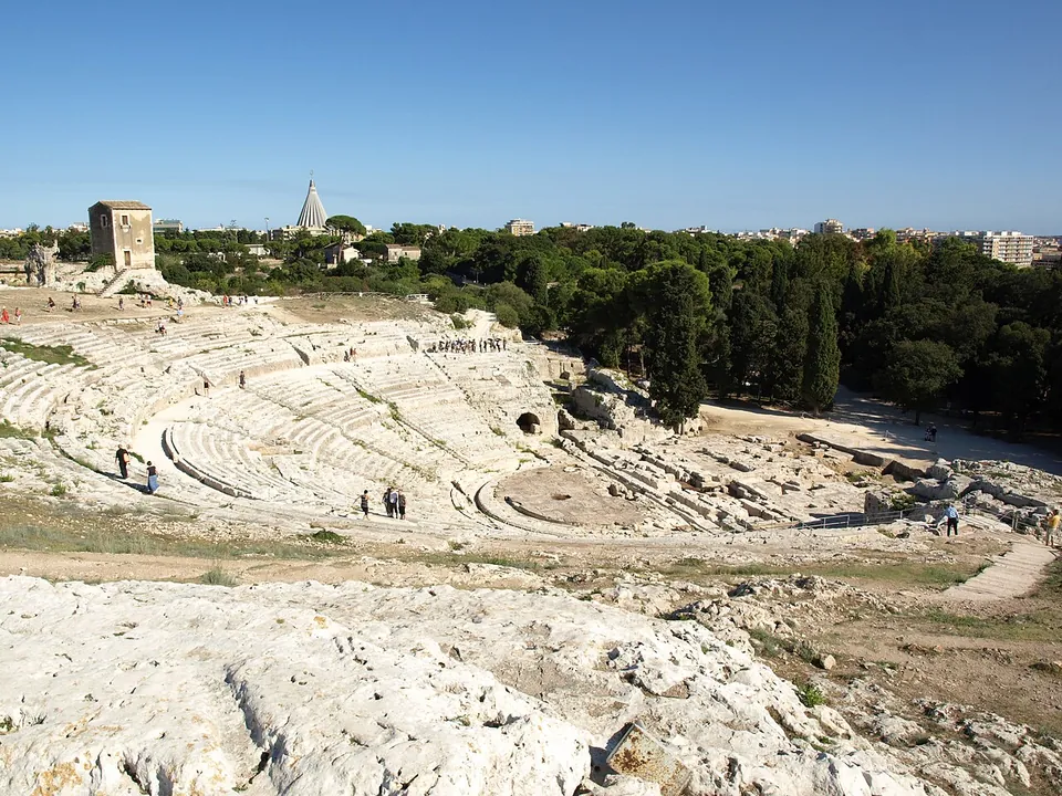 The great semicircle of pale stone seating cut into the hillside, divided into wedges by stairways, with a few visitors on the steps, a square stone tower on the rim at left and a tall conical modern church on the skyline.