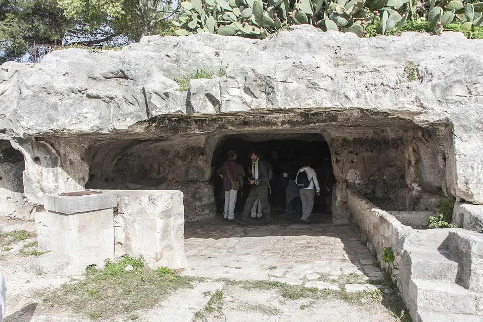 A wide, low opening cut into a pale limestone outcrop, with visitors standing inside the shadowed entrance, cut stone blocks and steps in front, and prickly pear growing on top of the rock.