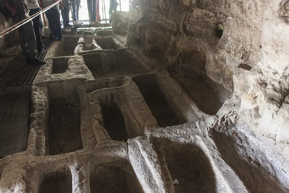Inside a rock-cut burial chamber, the floor is cut into rows of rectangular grave trenches separated by narrow ridges of stone, with a timber walkway and a rope barrier along one side and visitors' legs visible at the top.