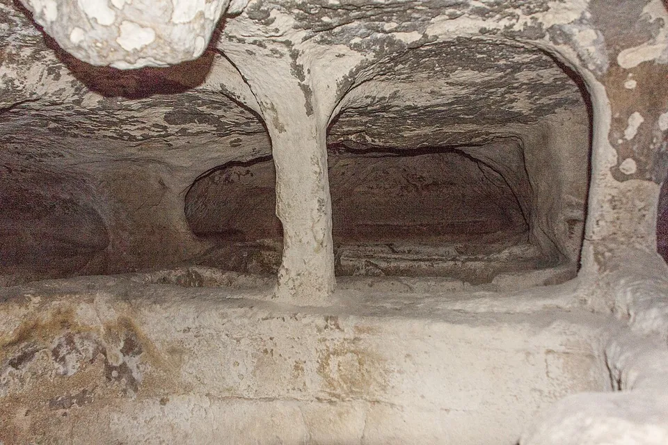 Two arched burial niches cut side by side into pale rock, divided by a slender rock-cut pillar, each niche holding a grave hollowed out of the stone, the arches edged with a reddish band.