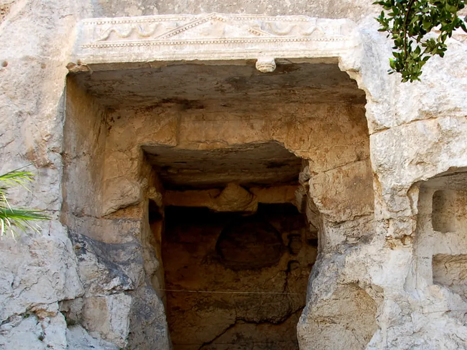 A doorway cut into a pale rock face, framed by a carved pediment with a low-relief frieze, opening into a dark rock-cut chamber.