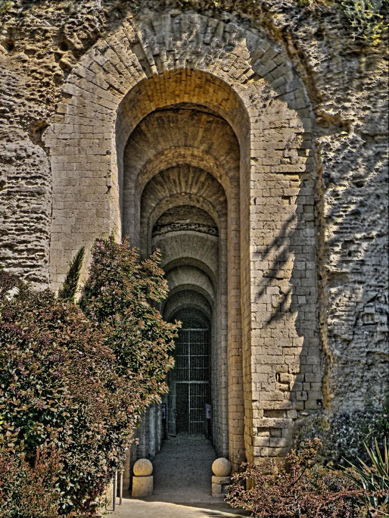 A tall arched opening faced with cut stone in a rough rock face, with a run of further arches receding into a dark tunnel behind it and shrubs on either side.