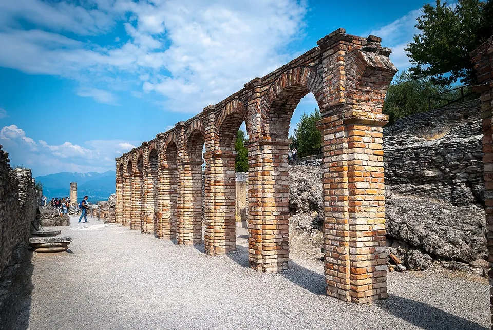 A row of brick piers joined by round brick arches runs beside a wide gravel path, with a wall of rough stone behind them and visitors further along.