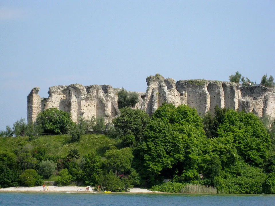 Seen from the water, a long ragged line of pale stone walls and piers stands on top of a wooded bank above a narrow beach on the lake shore.