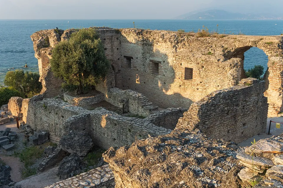 High walls of rubble stone with rows of small rectangular openings enclose sunken rooms, an olive tree growing inside one of them, and an arched opening at the right looks out over the lake.