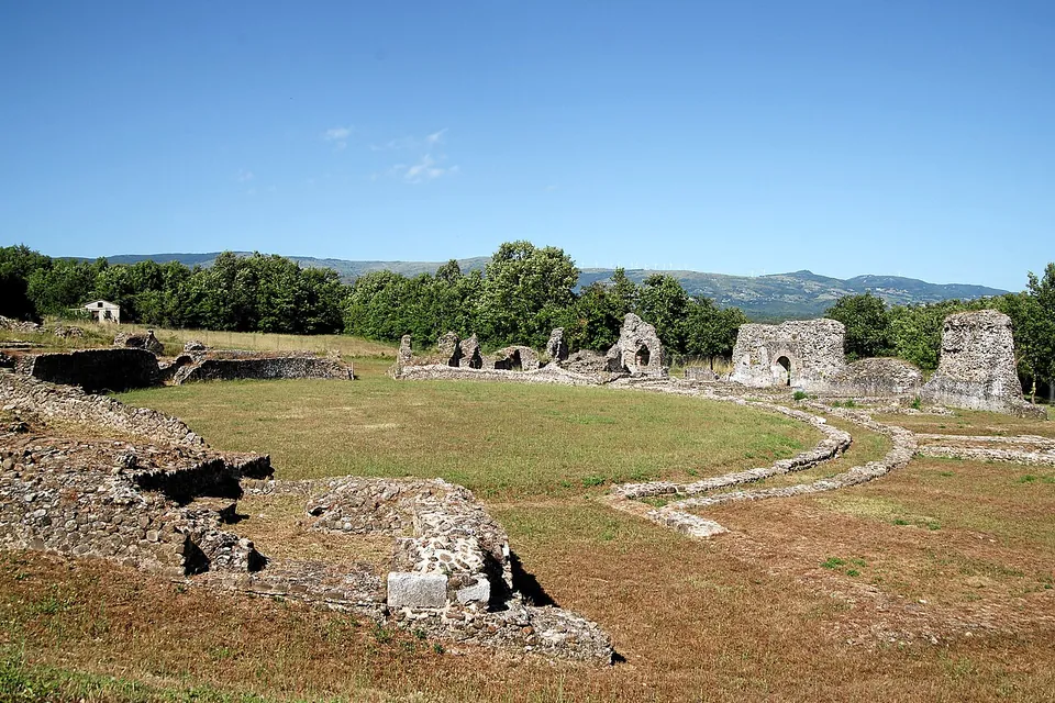 The grassed oval of an arena ringed by a low curving wall, with ruined stretches of the surrounding seating structure and arched passages standing on the far side against wooded hills.