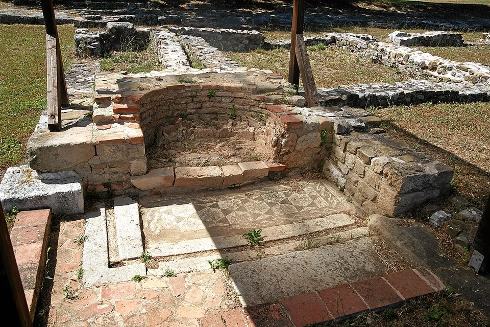 A small apse-shaped niche of stone and brick, with a mosaic floor of small patterned squares in front of it and low excavated walls beyond, under a timber support.