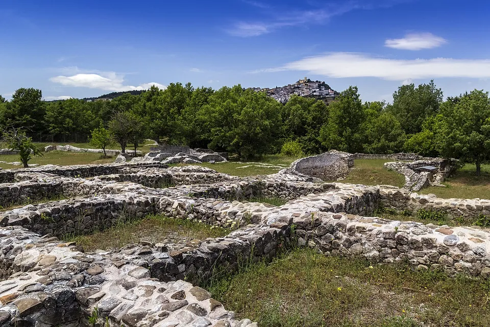 Low walls of rough grey stone laid out across grass among scattered trees, with a town of pale houses crowning a hill on the skyline behind.