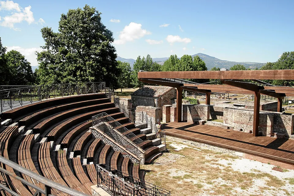 Curving tiers of theatre seating with modern railings and a metal walkway, facing a stage area under a timber frame, with trees and mountains behind.