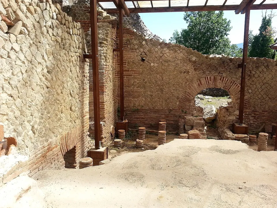 Short stacks of round bricks standing in rows on a floor, beneath walls of small square stones set in a diagonal net pattern with brick bands and a brick arch, under a modern timber roof.