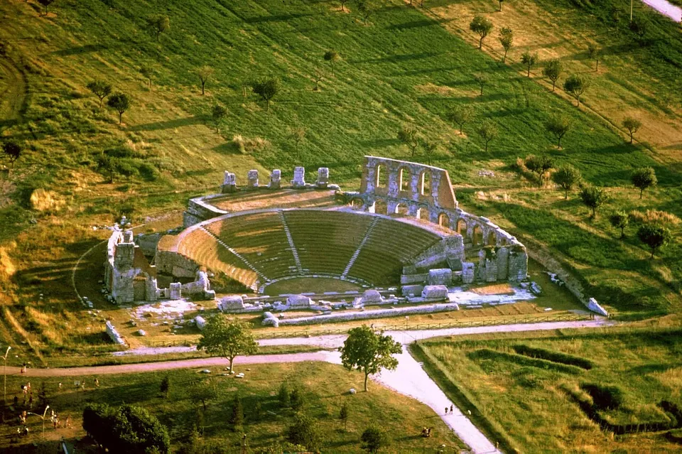 An aerial view of the theatre in a mown meadow - the semicircle of stepped seating, the ring of arches behind it with a taller section of two-storey arcade standing at one end, the stage foundations across the front, and gravel paths leading in past scattered trees.