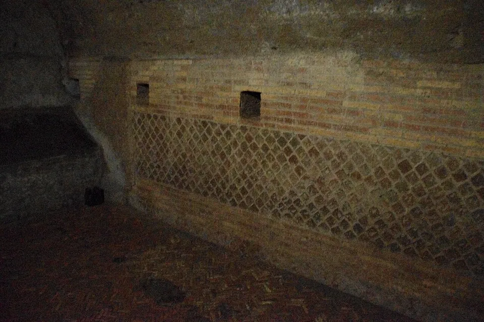 A dimly lit underground wall of narrow red bricks in horizontal courses over a band of small square stones set in a diagonal net pattern, with two small square holes in the brickwork and a brick floor.