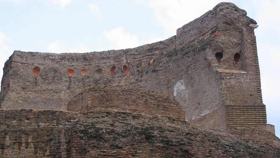The broken top of a brick and concrete wall in which a row of round clay vessel mouths is set, seen from below against the sky.