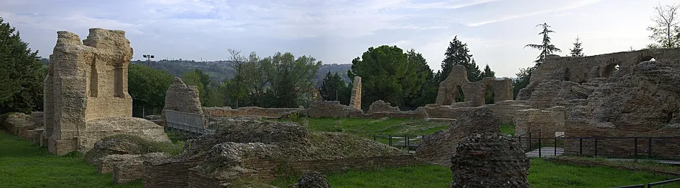 A wide panorama across mown grass scattered with low brick and stone ruins, a tall pier of masonry standing on the left and a long ruined wall with arched openings on the right, with trees and hills behind.