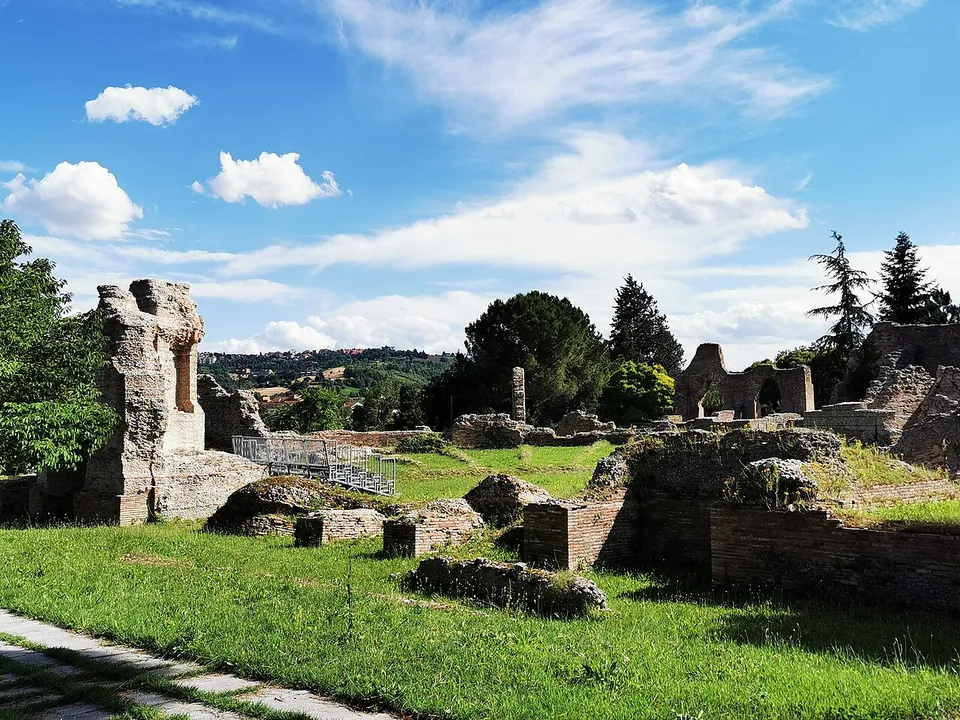 A paved path running past grass and low brick walls towards a tall ruined pier with a window-like opening, with more arched ruins among trees in the background.