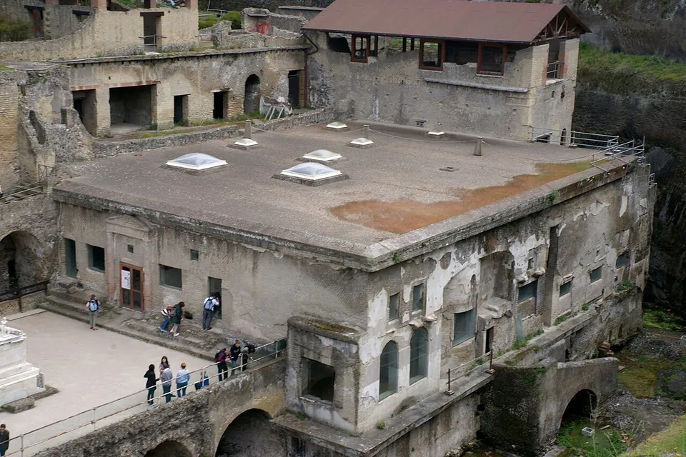 A large, almost intact Roman building with a flat roof pierced by skylights and walls with arched and square windows, seen from above, with visitors on the paved terrace beside it.