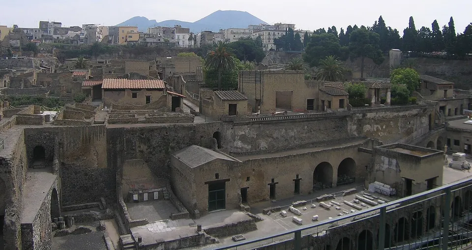 A view down into the excavated town from the modern street level, roofless and roofed Roman houses packed along narrow streets in a deep pit, with the apartment blocks of modern Ercolano above it and the cone of Vesuvius on the horizon.