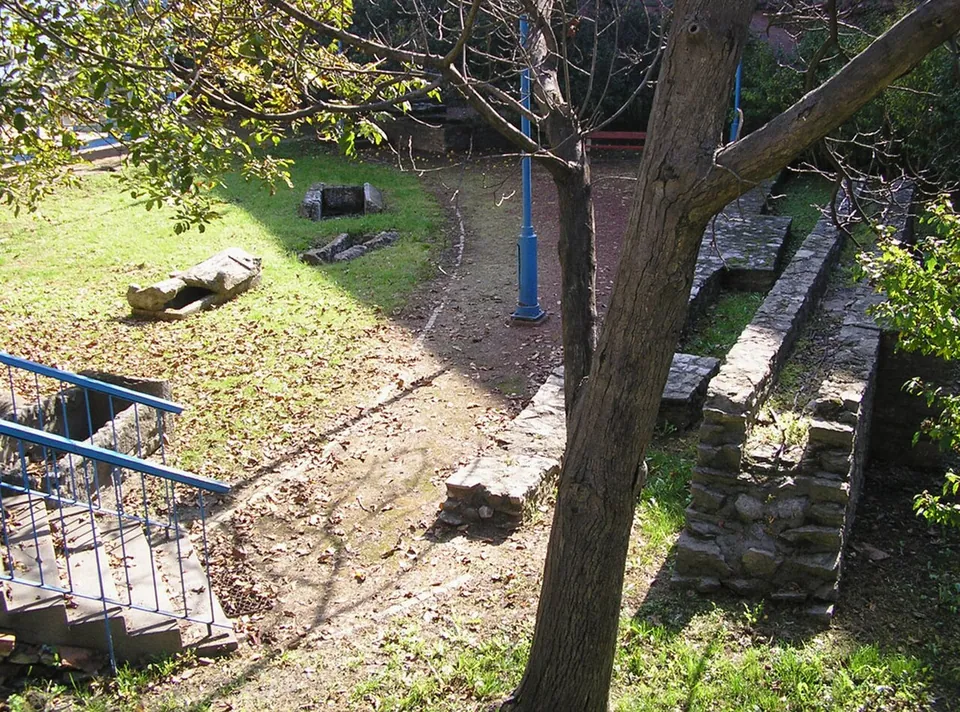 A garden with trees and a lawn, low stone walls of excavated rooms, two stone coffins lying on the grass and blue railings around a stair on the left.